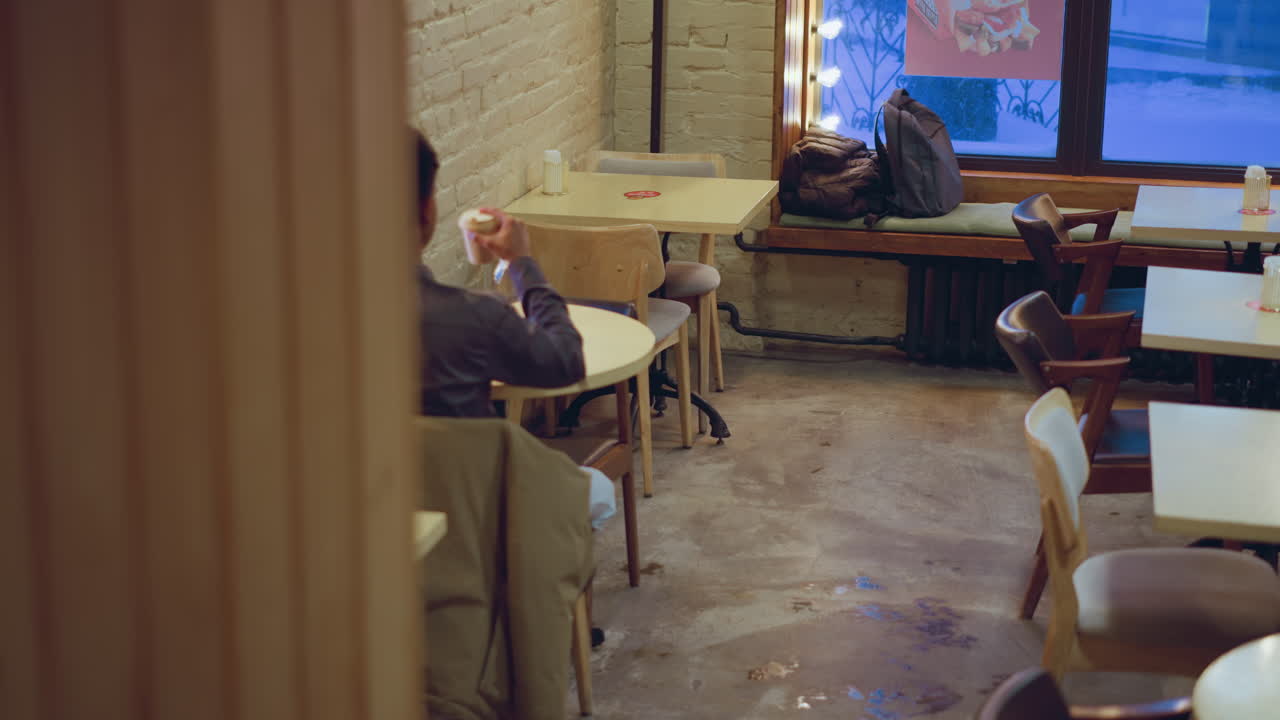 Student wearing formal shirt sits quietly inside cozy cafe holding takeaway coffee cup, facing softly lit window with glowing lights, surrounded by empty chairs, jackets, backpacks, and textured brick walls