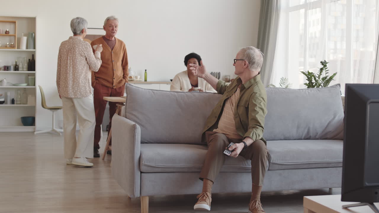 Wide shot of senior Caucasian man sitting on couch in living room, watching TV emotionally, calling for his friends they joining from background