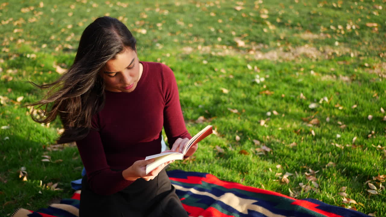 Close up of a young hispanic woman college student reading a book outdoors in the grass before class on campus