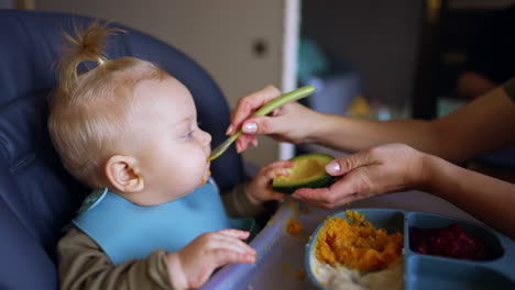 mom's hands hold a half of the avocado and a spoon. woman scrubs some fruit and gives it to the baby. close up.