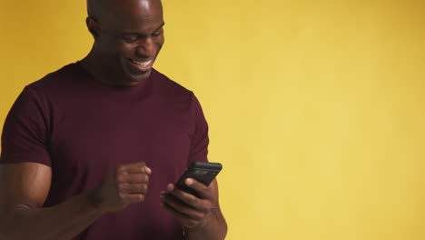 studio shot of excited mature man with mobile phone celebrating winning money against yellow background