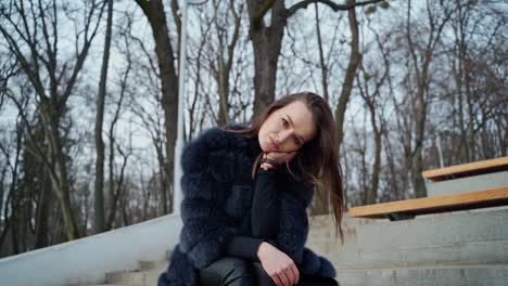 portrait of attractive model sitting in the park. beautiful young woman in black fur coat posing on camera outdoors.