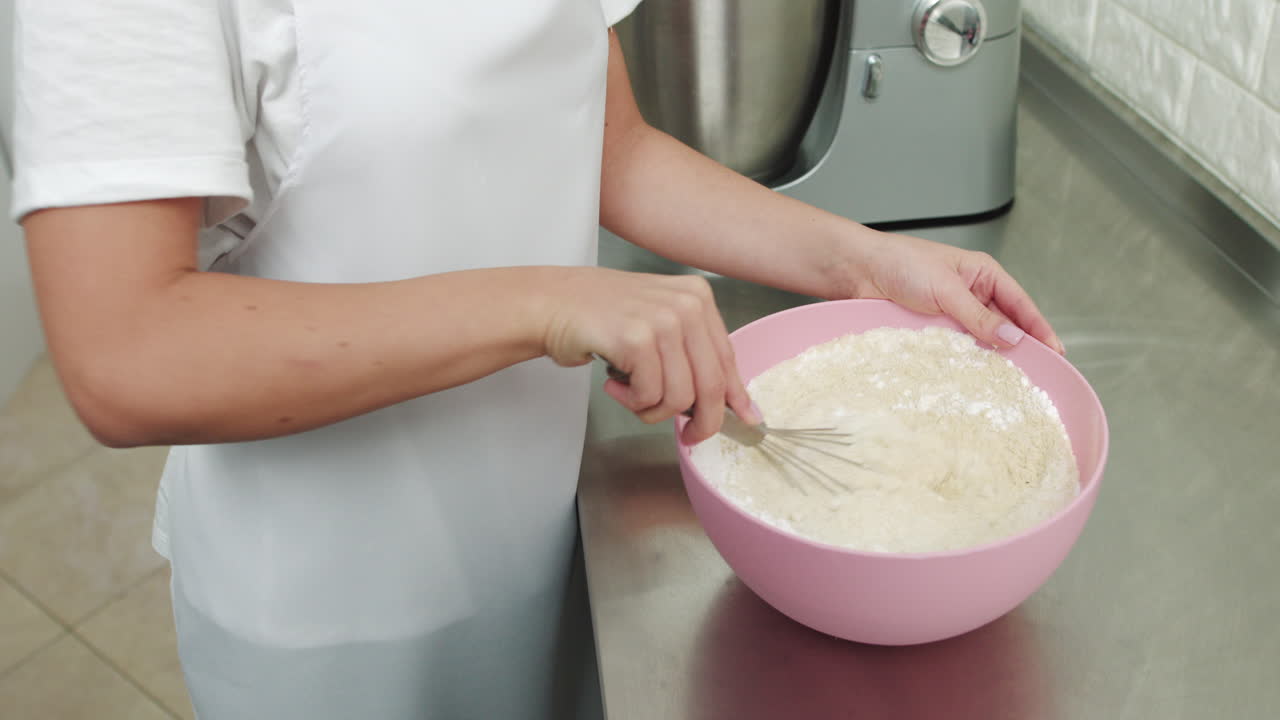 Chef mixing the flour in a pink bowl using a whisk while standing in light confectionery interior
