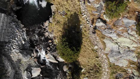 aerial: zenital shot of a river and two hikers walking a path in the mountains during late autumn or early winter