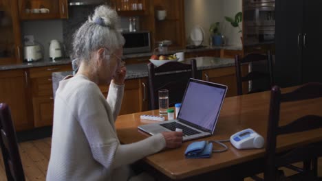 mujer mayor caucásica sosteniendo una medicación vacía mientras hace una videollamada en una computadora portátil en casa