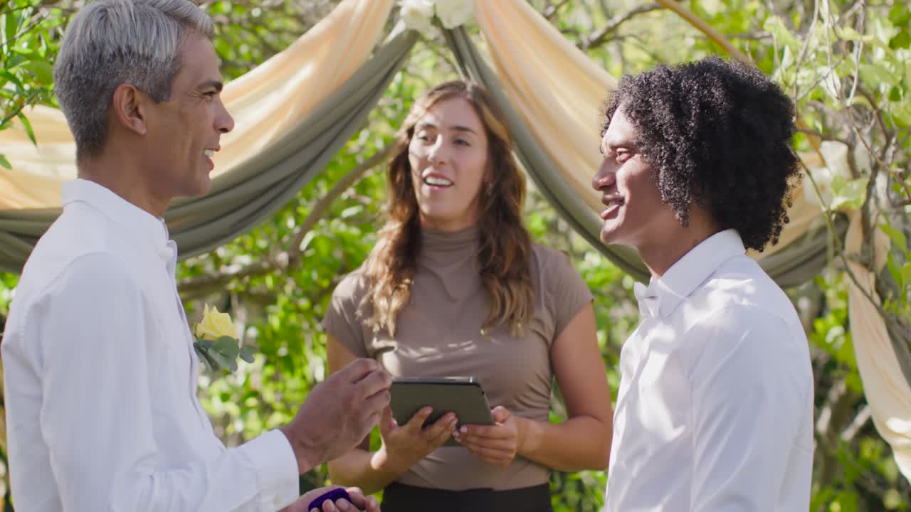 Male pair holding ring box, female officiant reading tablet, leading ring exchange at garden arch