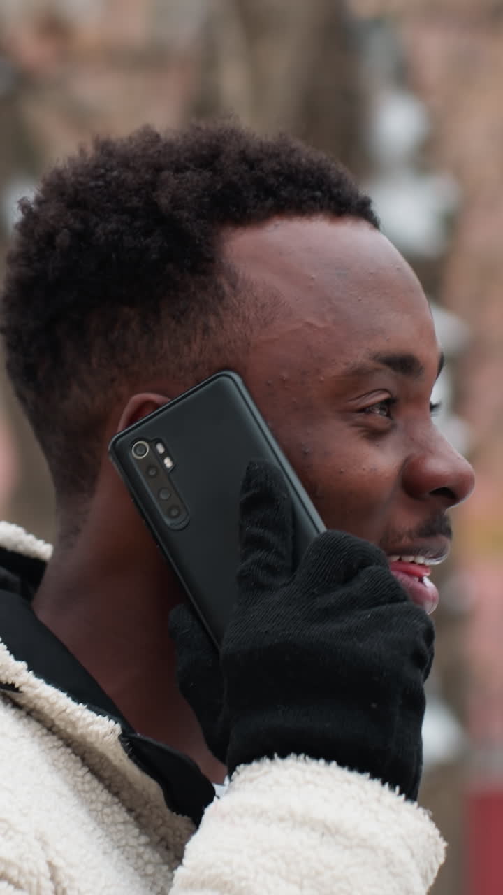 vista lateral de un hombre negro feliz conversando por teléfono, sonriendo en un entorno invernal al aire libre con árboles desnudos y edificios en el fondo, disfrutando de la conversación