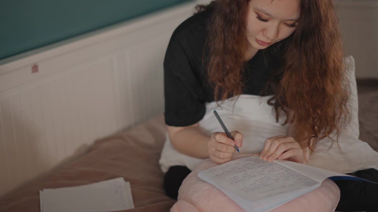 Giovane donna seduta sul letto in una camera da letto accogliente che scrive appunti su carta, chinata in avanti con concentrazione focalizzata, facendo una pausa tra le righe mentre riflette nella morbida luce del giorno, con elementi come donna, scrittura, letto, penna