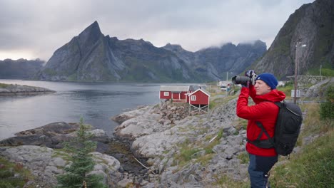 un fotógrafo masculino con una chaqueta roja está tomando una foto de las montañas y los fiordos con su lente telefoto en hamnøy, islas lofoten, noruega