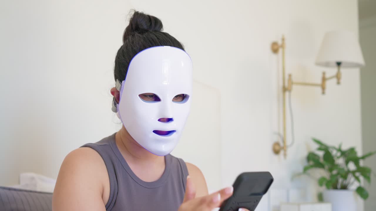 Eine Frau hält ihr Handy nah, scrollt, um Nachrichten zu prüfen, während sie eine weiße LED-Maske im Bett trägt.