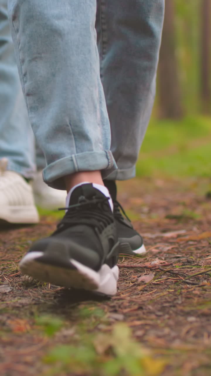 primer plano de dos damas caminando por un sendero del bosque, cada una con pantalones vaqueros azules, una con zapatillas blancas y la otra con negras, sus pasos se mueven en sincronía sobre un camino cubierto de cones de pino y hojas