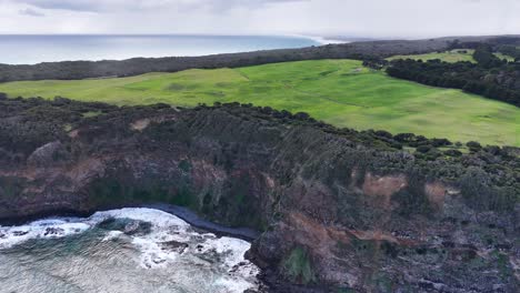 Drone pans over Cape Schanck cliffs, green fields, and crashing ocean waves under cloudy daylight