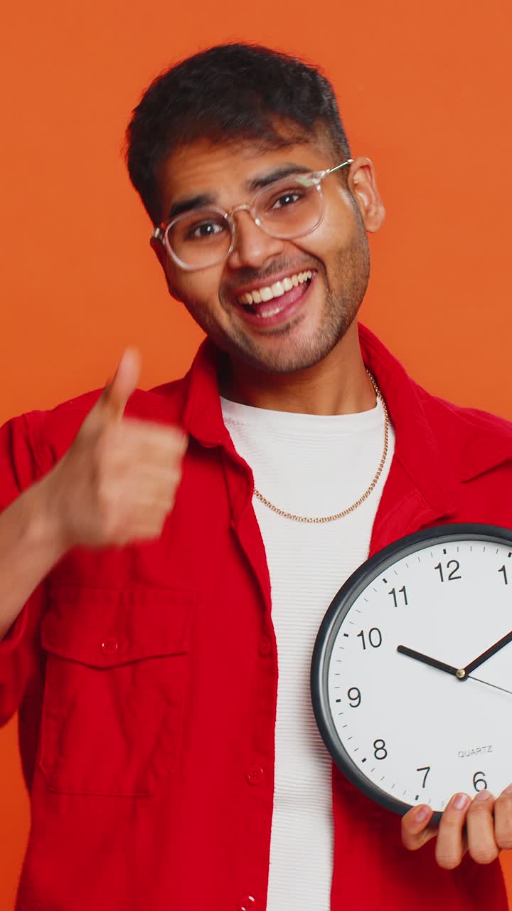 Indian young man showing time hour on wall office clock ok thumb up approve hurry up deadline