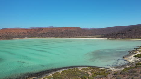 Filmische-Drohnenaufnahme-Vom-Strand-Von-Balandra,-Die-über-Rote-Hügel,-Türkisfarbenes-Wasser,-Weiße-Sandstrände-Und-Berge-Im-Hintergrund-Fliegt