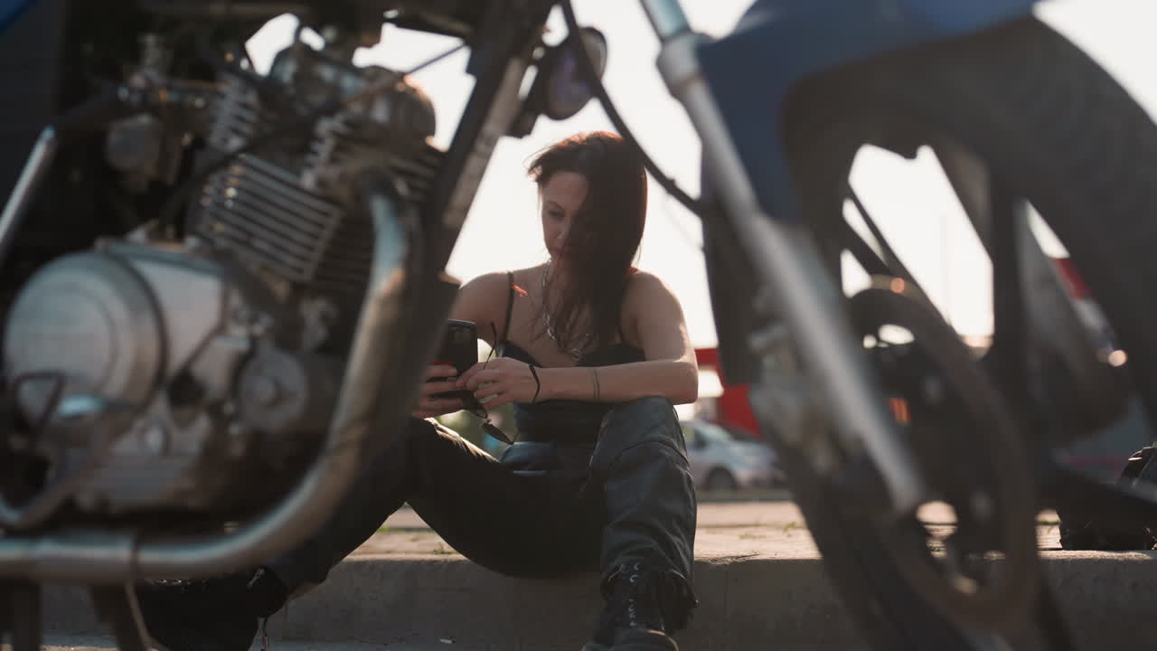 Close up of parked motorbike framing woman seated on pavement operating phone with helmet placed by side slight blur background of parked car and building under daylight