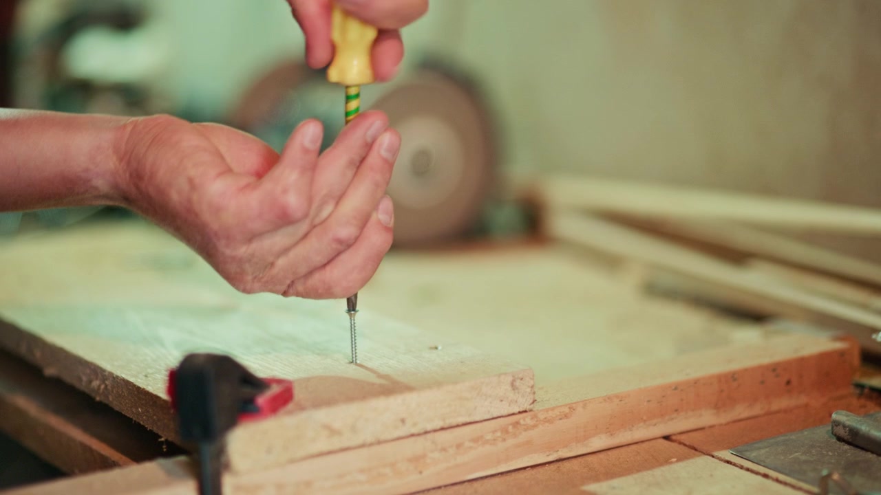 Close-up of a craftsman using a manual screwdriver to fasten a screw into a wooden plank in a workshop. Authentic handmade DIY woodworking footage for craft and maker projects