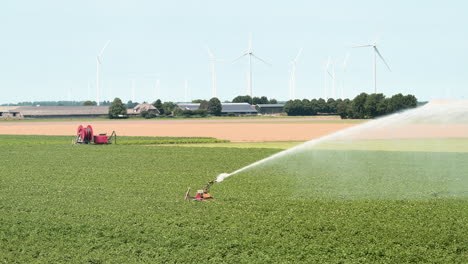 Sistema-De-Riego-Agrícola-Rociando-Agua-Sobre-Cultivos,-Aerogeneradores-En-Segundo-Plano,-Cámara-Lenta-1