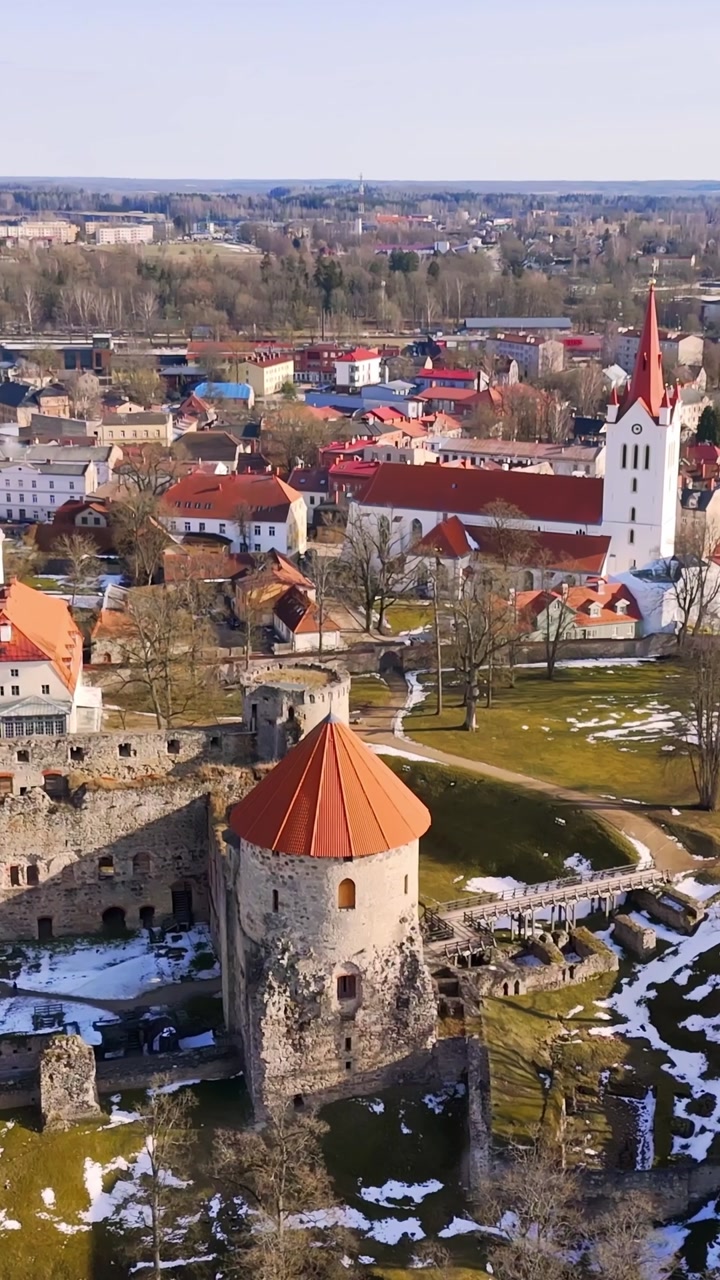 Vertical aerial view of Cesis Castle and St. John’s Church, capturing medieval towers, old town rooftops, and early spring snow patches in bright daylight.