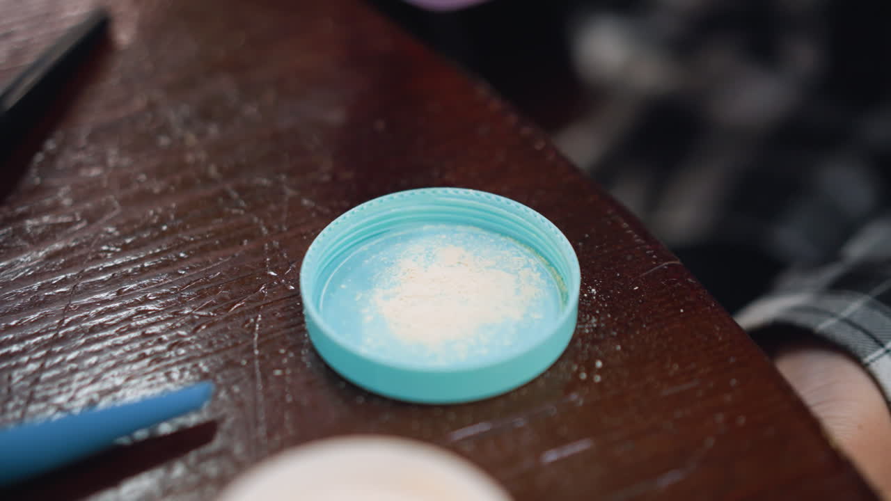 primer plano de la mano de la dama sosteniendo una esponja de maquillaje púrpura, tocando en el polvo en la cubierta en la mesa, esmalte de uñas visible en los dedos, configuración de rutina de belleza con herramientas de maquilaje y polvo en superficie