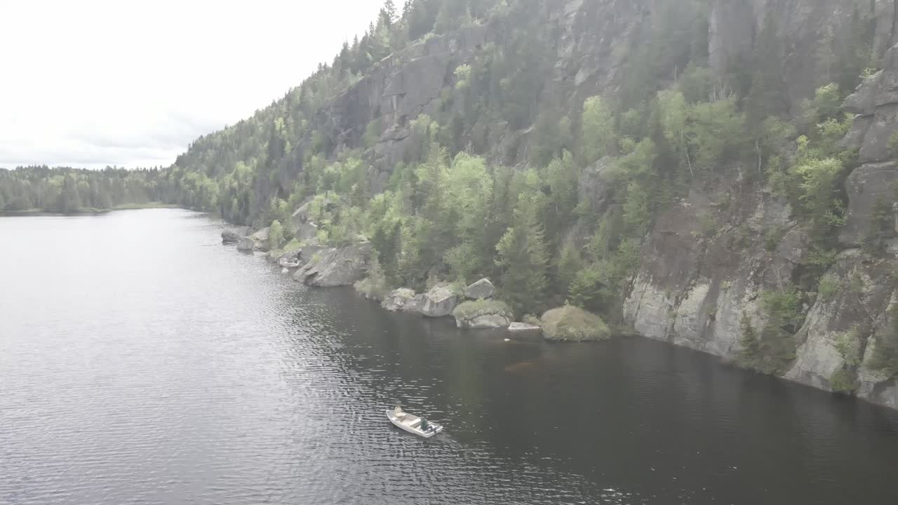 Sweeping drone descends to reveal fisherman navigating near towering rock face on tranquil forest lake.