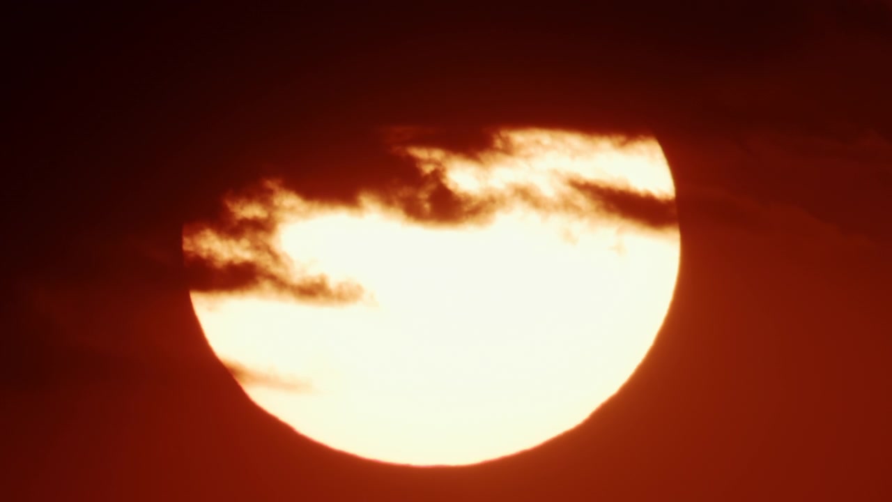 分厚い暗い雲の帯の向こうに太陽がゆっくりと沈み、その下に輝く水平線が見えるワイドショット。
