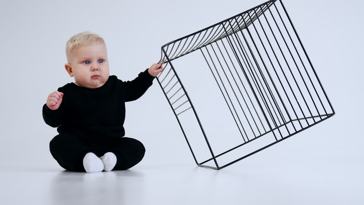 Little cute blond baby in black suit and white socks sits on the floor. Kid grabs and pulls a cage near him closer but adult stops him. White backdrop.