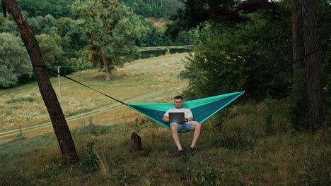caucasian man sits swaying in the hammock. freelancer doing remote job on his laptop in the nature.