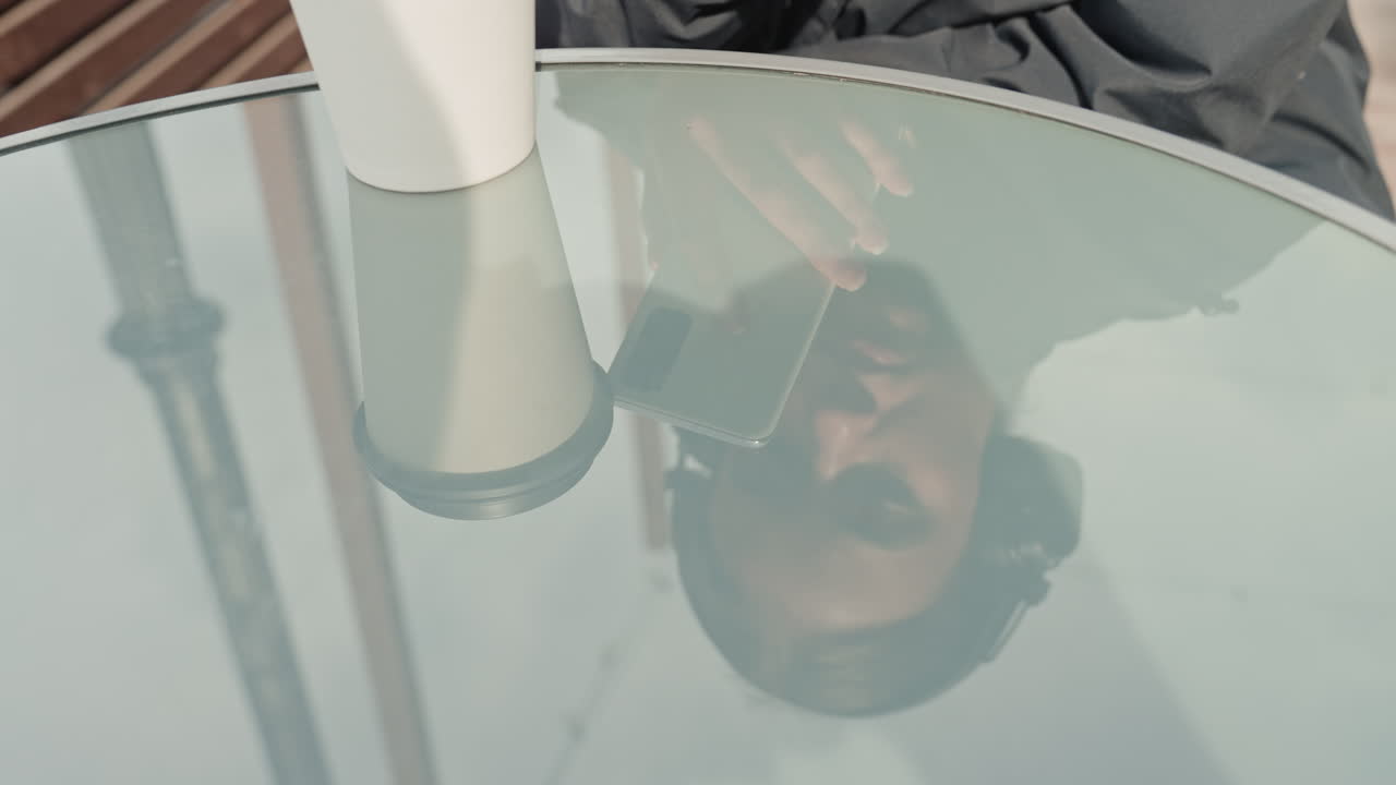 Donna riflessa sul tavolo di vetro che tiene in mano il telefono e sorseggia il caffè, controllando i messaggi. Terrazza di un bar all'aperto sotto una tenue luce mattutina, atmosfera pensierosa, primo piano del riflesso con profondità di campo ridotta, giacca casual