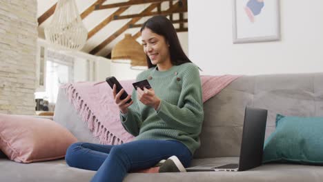 biracial woman using smartphone and holding credit card in living room