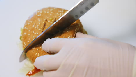 hands in gloves of a cook making burger. chef cutting ready hamburger into halves with a sharp knife on table. close-up.