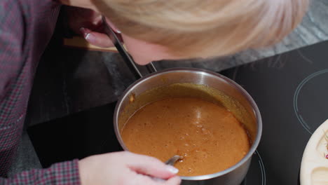 close-up of woman cooking, leaning down to smell the food while holding the pot handle on an electric stovetop, the bubbling dish simmers in a modern kitchen setting with a sleek countertop