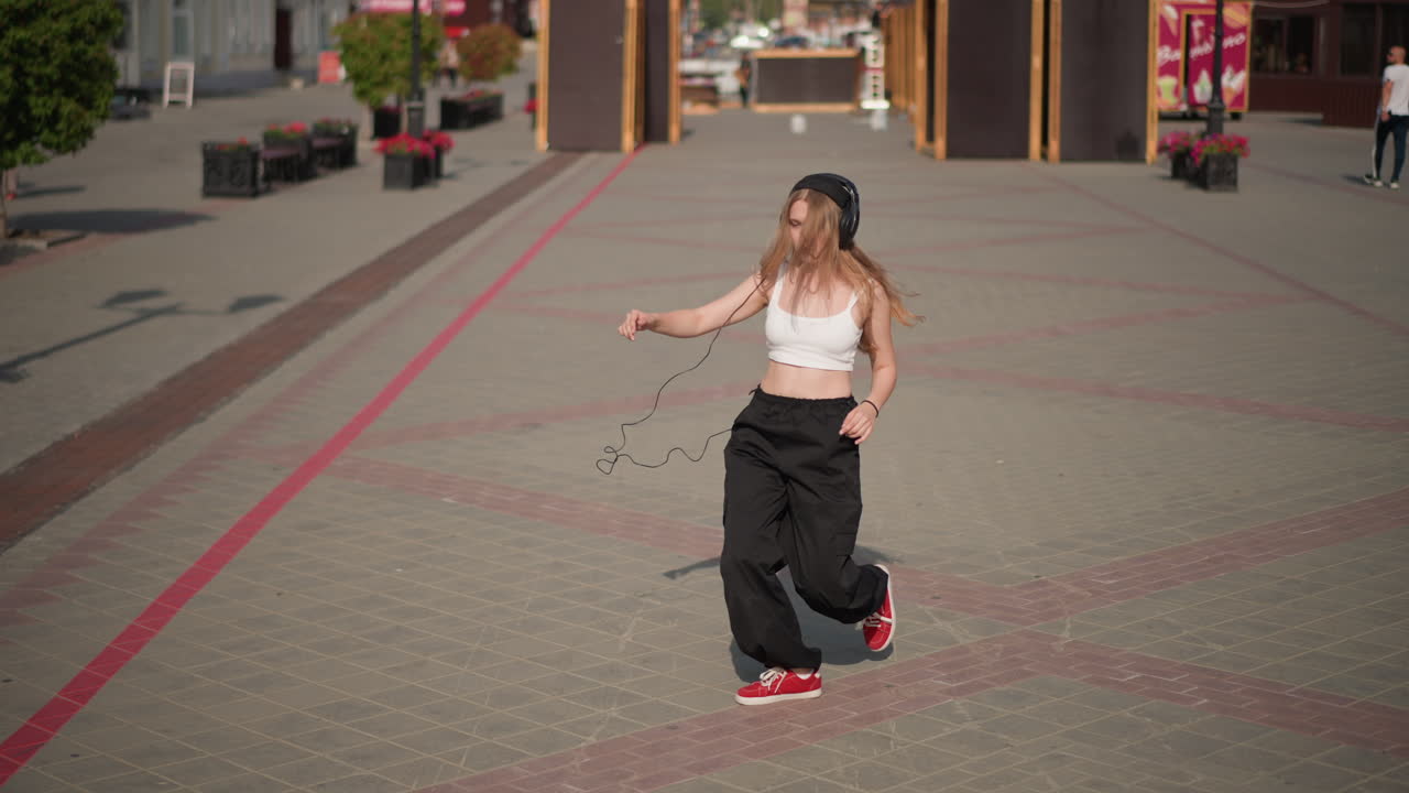 Giovane donna che balla per strada in una piazza vuota, indossa un top corto e pantaloni neri larghi con scarpe da ginnastica e berretto rossi, esegue energici movimenti freestyle con rotazioni, scivolate, corse e pose, espressione sicura di sé