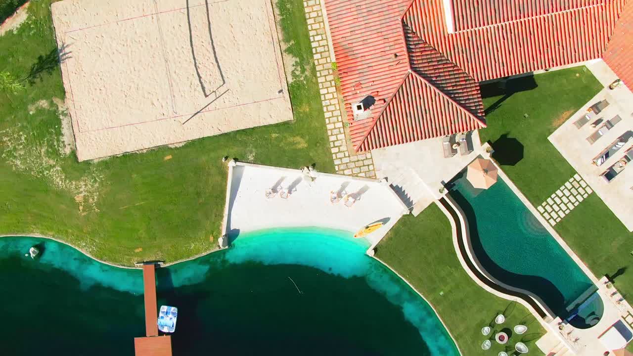 Large luxury holiday home with large green lawns, a swimming pool and white sandy beach at the clear blue water with relaxing chairs. Top down rotating shot