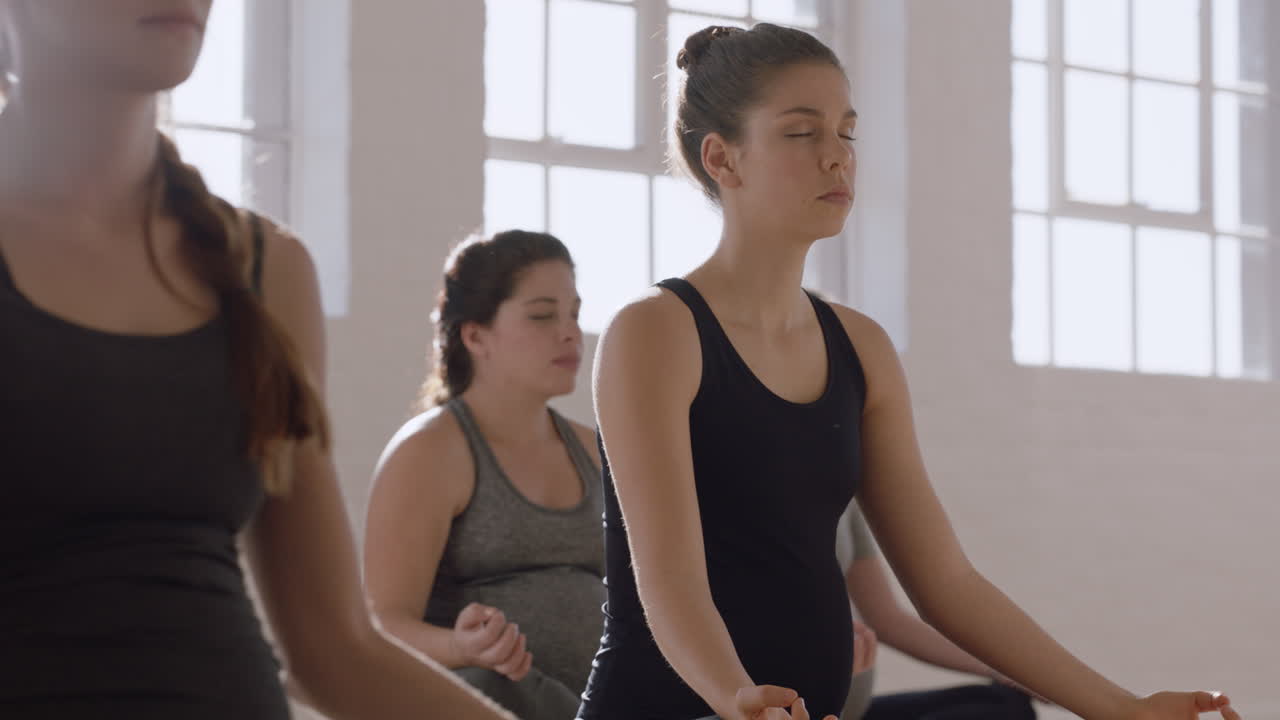 joven mujer caucásica embarazada en clase de yoga practicando postura de loto disfrutando de la práctica de meditación en grupo relajándose en el gimnasio al amanecer.