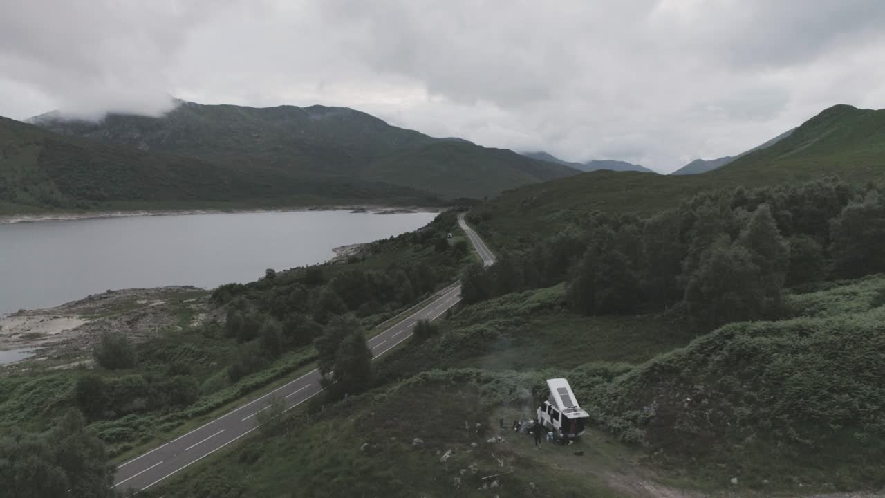 Static mid drone shot showing a campervan wild camping with BBQ smoke and Loch Beag and cloudy mountains in the background in Scottish Highlands