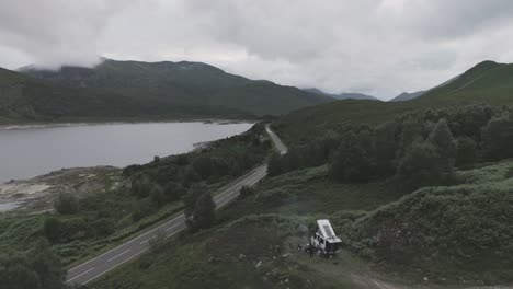 static mid drone shot showing a campervan wild camping with bbq smoke and loch beag and cloudy mountains in the background in scottish highlands