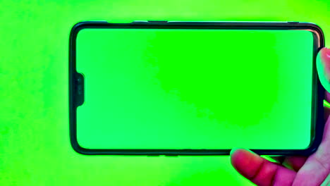 mano cercana de un hombre asiático que sostiene un teléfono inteligente moderno contra un fondo de pantalla verde con pantalla verde