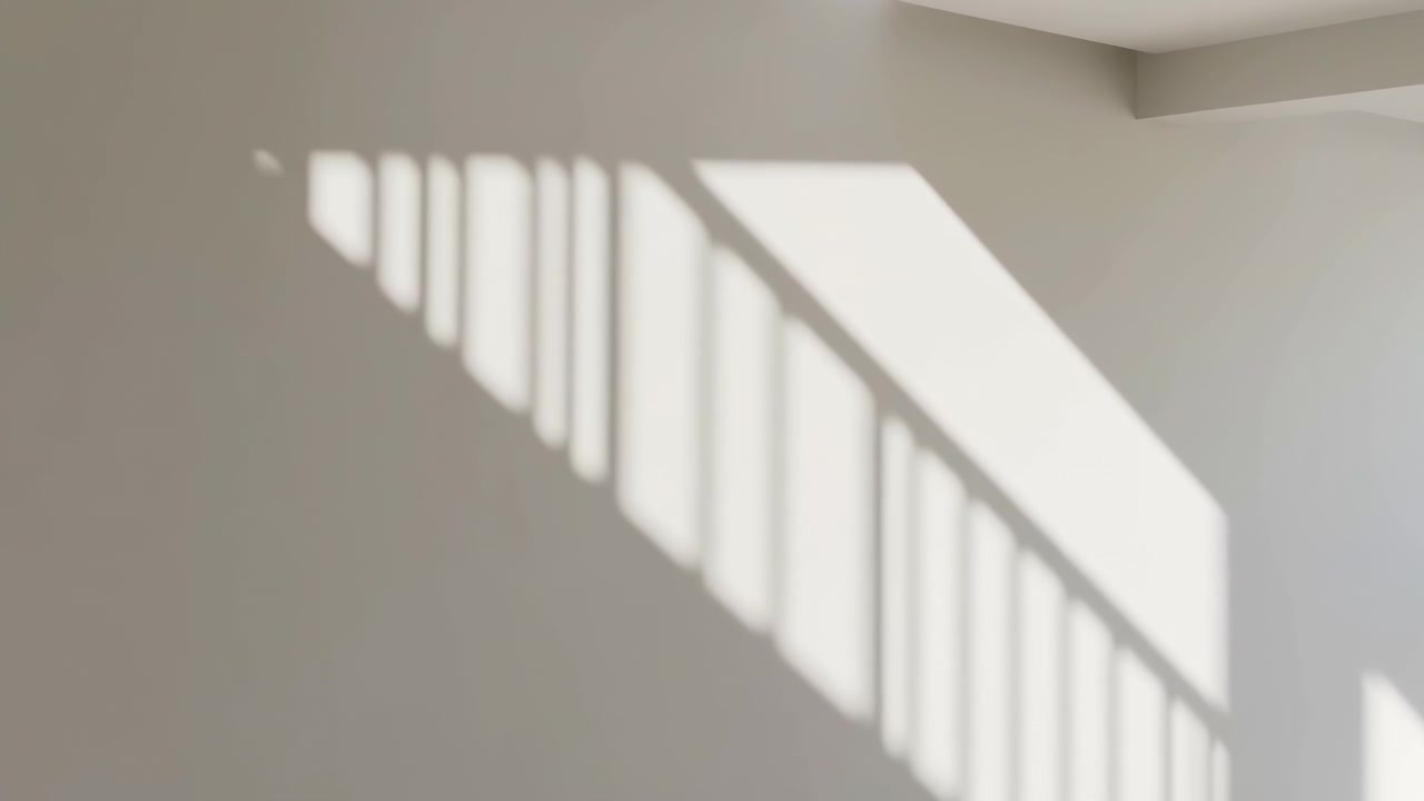 Bright diagonal sunlight and soft window frame shadows fall across a clean beige wall in a minimalist room, creating a calm abstract pattern of light and shade