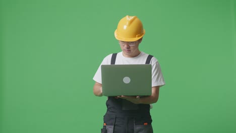 asian man worker wearing goggles and safety helmet using a laptop while standing in the green screen background studio