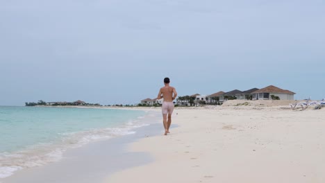 Joven-Blanco-En-Topless-Corre-En-La-Orilla-De-La-Playa-En-Bimini,-Bahamas-A-Lo-Largo-De-Algunas-Villas-Nuevas