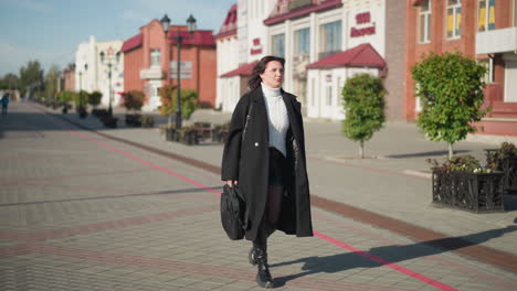 business lady going to her office in black coat, and black boots on paved urban street during sunny day, holding black handbag with someone blur in background, surrounded by modern buildings