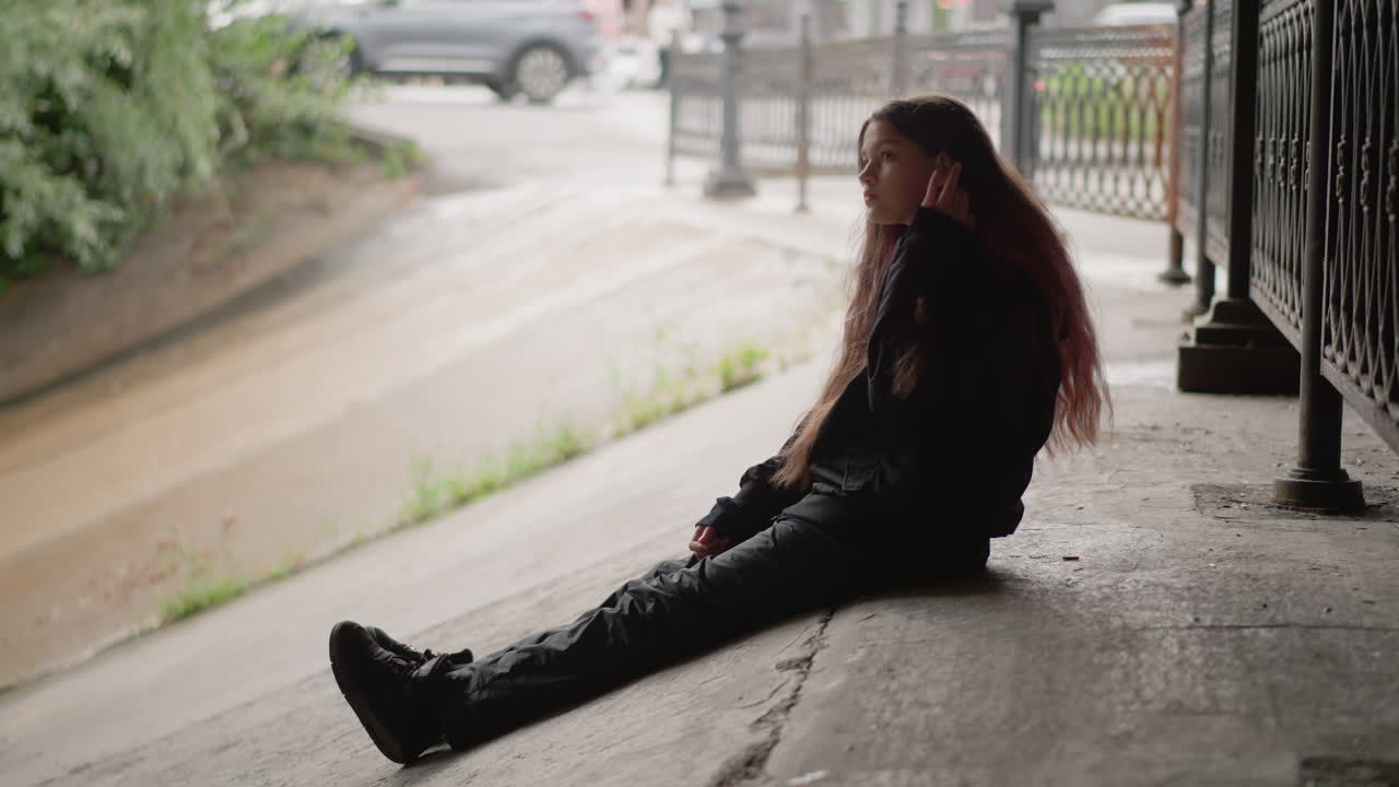 Mulher sentada no pavimento molhado pela chuva, cabelo comprido húmido, botas perto do reflexo de uma poça, rua urbana nublada ao fundo, grade de ferro emoldurando a cena, olhar contemplativo, tons cinematográficos suaves, nostálgico