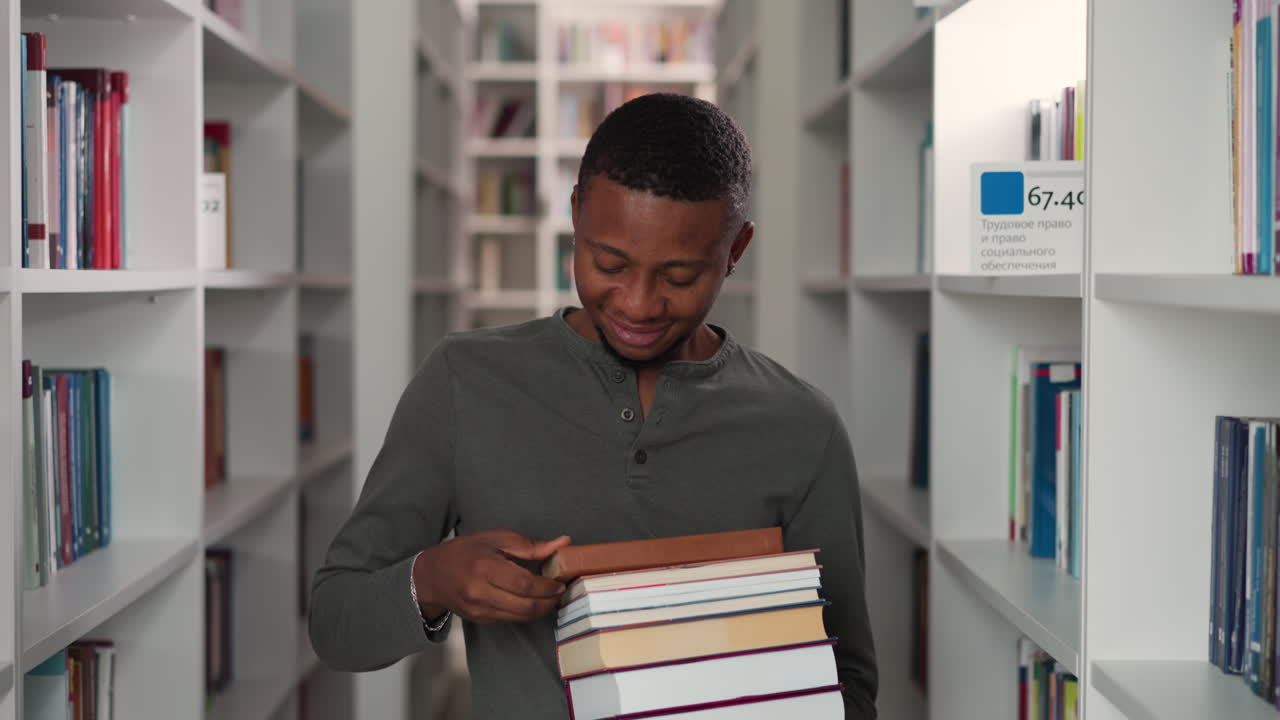 Smiling man holds books in city public library. African American reader carries literature stack between bookshelves. Black guy with chosen novels
