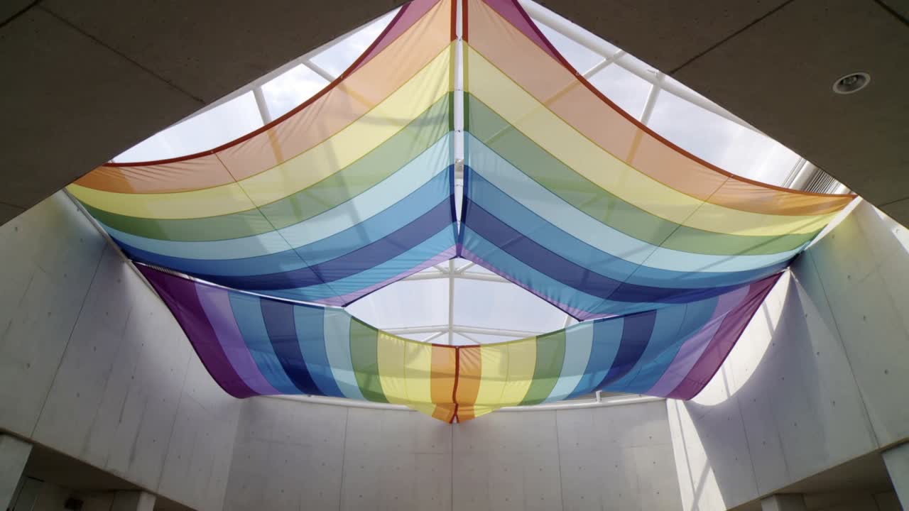 Tessuto a due strati a strisce arcobaleno che ondeggia e si increspa per la brezza sotto un lucernario, proiettando ombre.