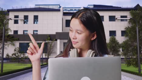 close up of asian teen girl student with a backpack use laptop and pointing to side while standing in front of a school building