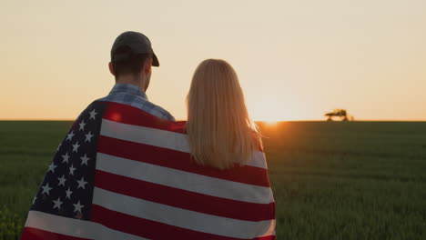 una pareja de agricultores con una bandera estadounidense en los hombros miran un campo de trigo donde un tractor está trabajando en la distancia.