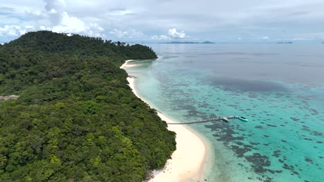 White-sandy-strip-of-beach-and-thin-pier-stretch-out-over-clear-blue-water-and-reef,-Ko-Rok-Thailand