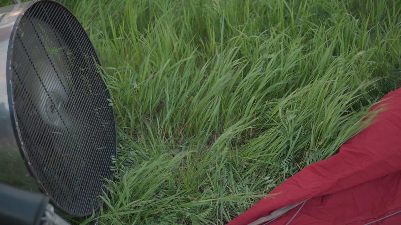 high speed hot air balloon fan engine rotating vigorously blowing wind through vibrant grass blades causing dynamic sway while red balloon fabric lies on lush green field under soft cloudy sky