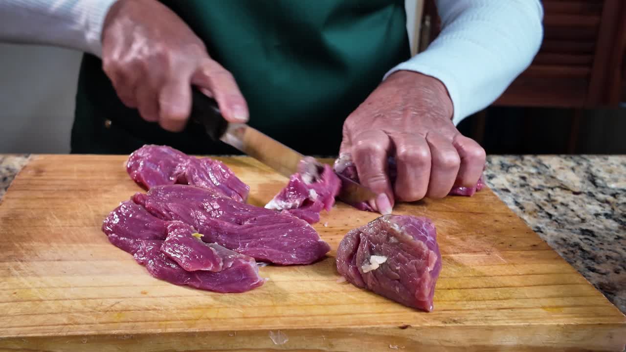 Wanita lansia memotong daging mentah di meja dapur dengan fokus dan hati-hati