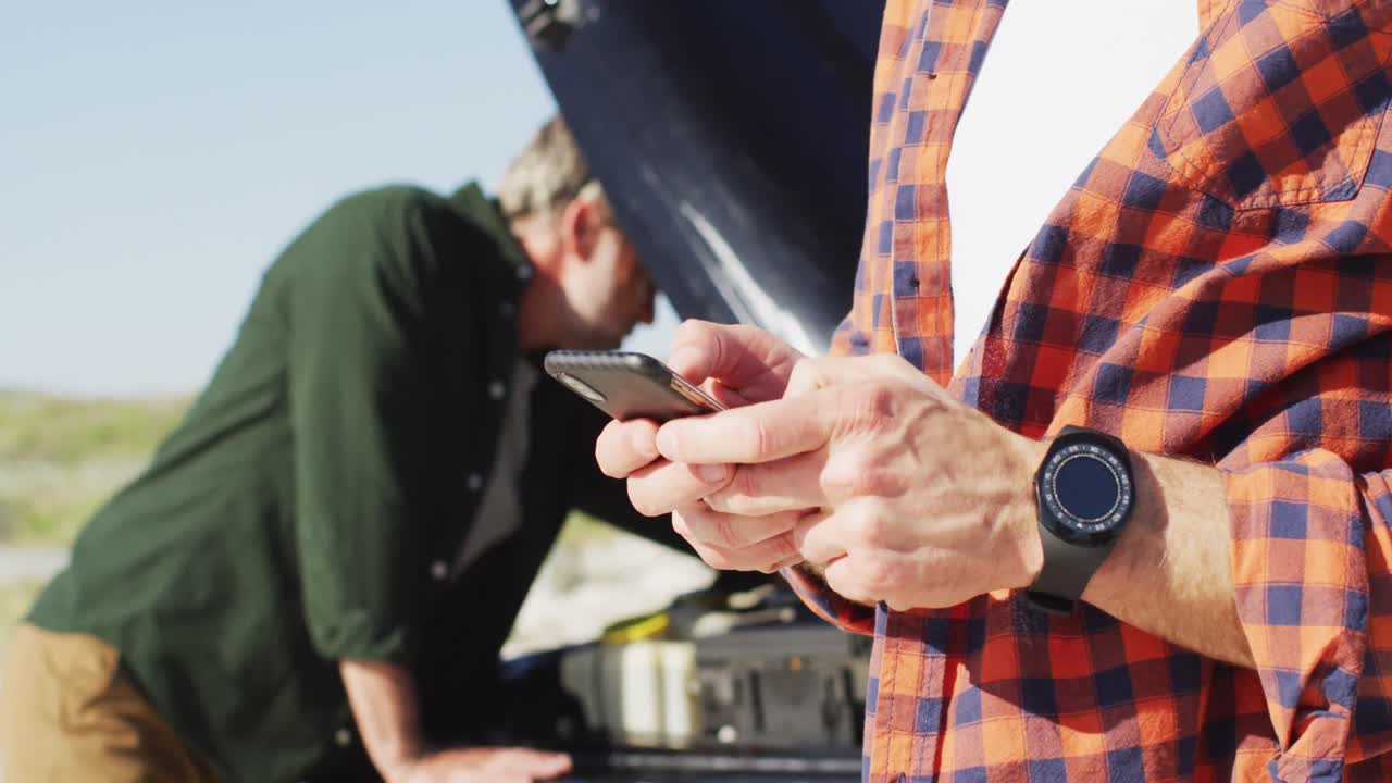 스트레스를 받은 백인 동성 남성 커플이 해변에서 스마트폰을 사용하여 고장난 차  ⁇ 에 서 있습니다.